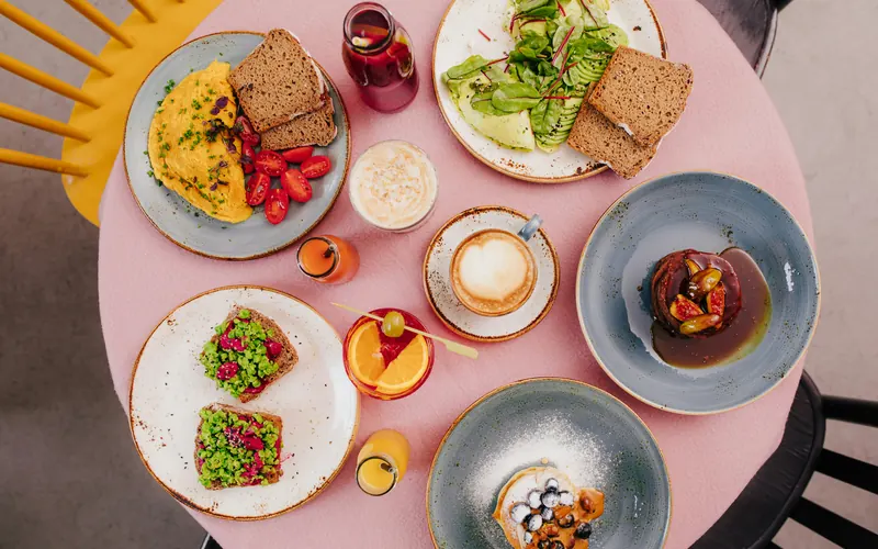 A pink brunch table seen from above with an omelette, avocado toast, pancakes and a cappuccino