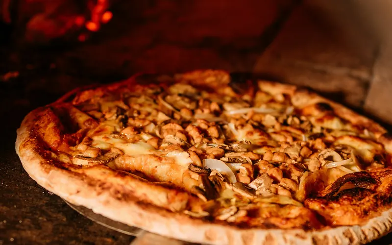 A pizza on a peel in front of the glowing fire of a wood-fired oven